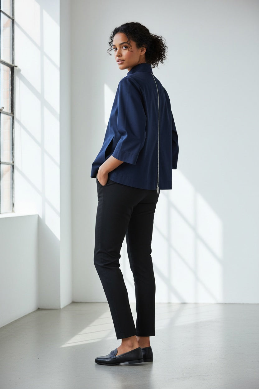 Standing against a light-colored wall with intricate molding, a woman with long hair reminiscent of Xena is turned away from the camera, showcasing the HINSON WU - XENA ZIP BACK SHIRT in navy blue. This A-line shaped top features an eye-catching gold tone zipper that extends down her back, perfectly complemented by elegant black pants.