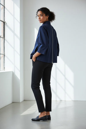 Standing against a light-colored wall with intricate molding, a woman with long hair reminiscent of Xena is turned away from the camera, showcasing the HINSON WU - XENA ZIP BACK SHIRT in navy blue. This A-line shaped top features an eye-catching gold tone zipper that extends down her back, perfectly complemented by elegant black pants.
