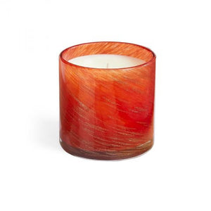 A LAFCO ABSINTHE PLUM HOLIDAY CANDLE, featuring an orange and red swirled hand-blown glass holder with a white candle inside. The design boasts a warm, fiery pattern reminiscent of swirling flames and exudes subtle hints of sandalwood.