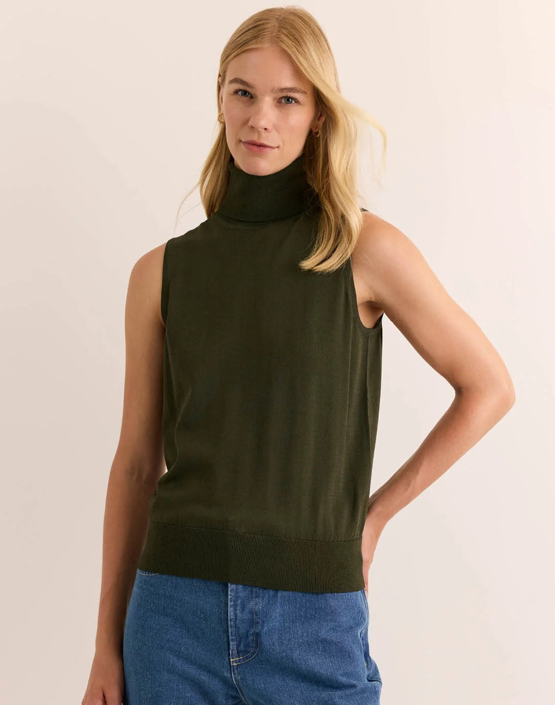 A woman with long blonde hair wears the HINSON WU - ASHLEY Sleeveless Turtleneck Knit Tank in Oregano, crafted from lightweight Verona Knit yarn, paired with blue jeans. She stands against a plain light background with one hand resting on her hip.