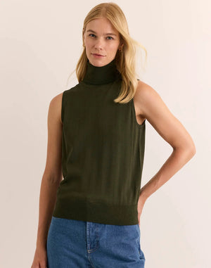 A woman with long blonde hair wears the HINSON WU - ASHLEY Sleeveless Turtleneck Knit Tank in Oregano, crafted from lightweight Verona Knit yarn, paired with blue jeans. She stands against a plain light background with one hand resting on her hip.