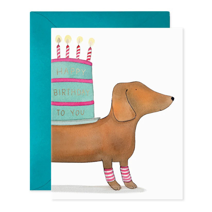 The E. FRANCES PAPER - SLINKY DOG BIRTHDAY GREETING CARD features a whimsical watercolor of a dachshund in pink and white socks carrying a cake with "Happy Birthday to You" on its back, complemented by a charming blue envelope background.