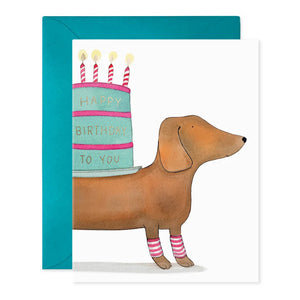 The E. FRANCES PAPER - SLINKY DOG BIRTHDAY GREETING CARD features a whimsical watercolor of a dachshund in pink and white socks carrying a cake with "Happy Birthday to You" on its back, complemented by a charming blue envelope background.