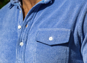 A person in a periwinkle Criquet Terrycloth Players Shirt and white shorts stands facing away from the camera against a plain beige background.