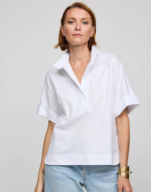 A woman with short light brown hair is seen standing against a plain light background, wearing the HINSON WU - CARA Elbow Sleeve Shirt in Stretch Polished Cotton White by HINSON WU. The oversized, loose-fit shirt features a polished cotton fabric with a collared neckline and elbow-length sleeves, and is paired with light blue jeans, large hoop earrings, and a gold bracelet. The machine-washable shirt offers effortless care.
