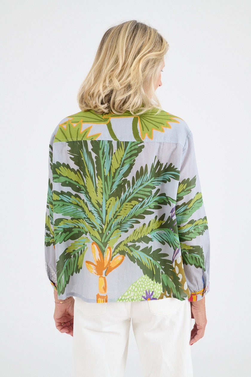 A woman with shoulder-length blonde hair wears the STORIATIPIC - SUZY CALI BLOUSE by STORIATIPIC, a light gray cotton top with a colorful palm tree print, paired with white pants and smiling gently against a plain white background.