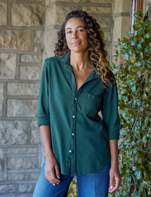 A woman with curly hair, smiling softly by a window indoors with one hand in her pocket, wears blue jeans and the FRANK & EILEEN Waverley Button Up Tee in Vintage Jersey Pine.