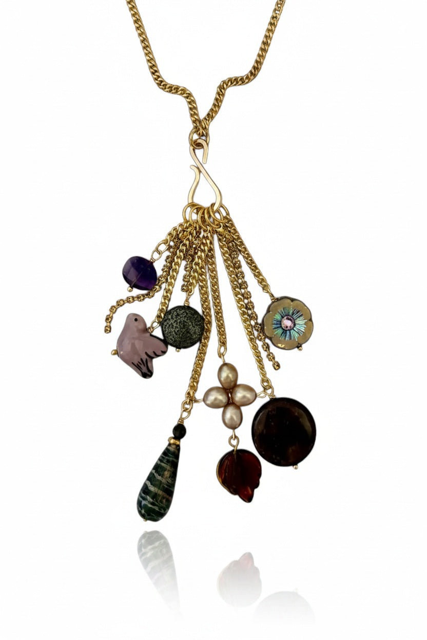 The SANDRINE FRANCE STUDIO - BORA PENDANT NECKLACE by SANDRINE FRANCE STUDIO features multiple charms—including colorful beads, pearls, a flower, a purple crystal, a teardrop stone, and other decorative elements—all artfully clustered on hanging gold chains to create a unique and eye-catching statement necklace.