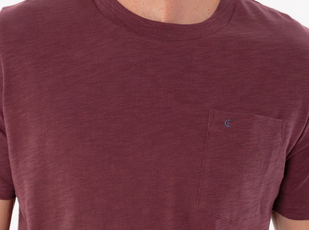 A person wearing the CRIQUET Slub Cotton Short Sleeve Pocket T-Shirt in Jam Band, featuring a small chest pocket and a subtle blue logo. The blurred greenery and wood background perfectly complement this vintage-inspired CRIQUET shirt, creating an effortlessly classic look.