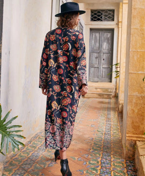 The MOMONI - FELICIEN DRESS by MOMONI is a long-sleeved, ankle-length black crepe de chine dress with a vibrant floral and paisley print in red, orange, and green, finished with patterned borders at the hem and cuffs.