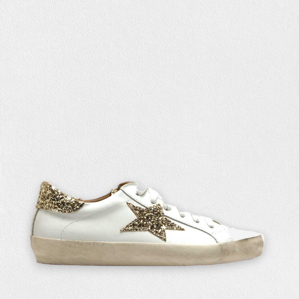 The MICHELE LOPRIORE - GEMMA SNEAKER WITH PLATINUM STAR features a white design with a worn sole. Adorning the side is a glittery platinum star, and the heel tab also sparkles with platinum glitter. Crafted from soft leather, this luxurious sneaker is showcased against a plain white textured background.