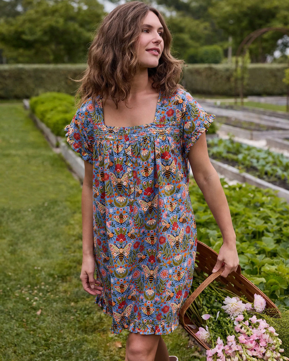 A woman with long blonde hair wears the PRINTFRESH Bee Tapestry Pinktuck Nightgown in Delphinium Blue, standing against a plain light-colored wall and smiling slightly.