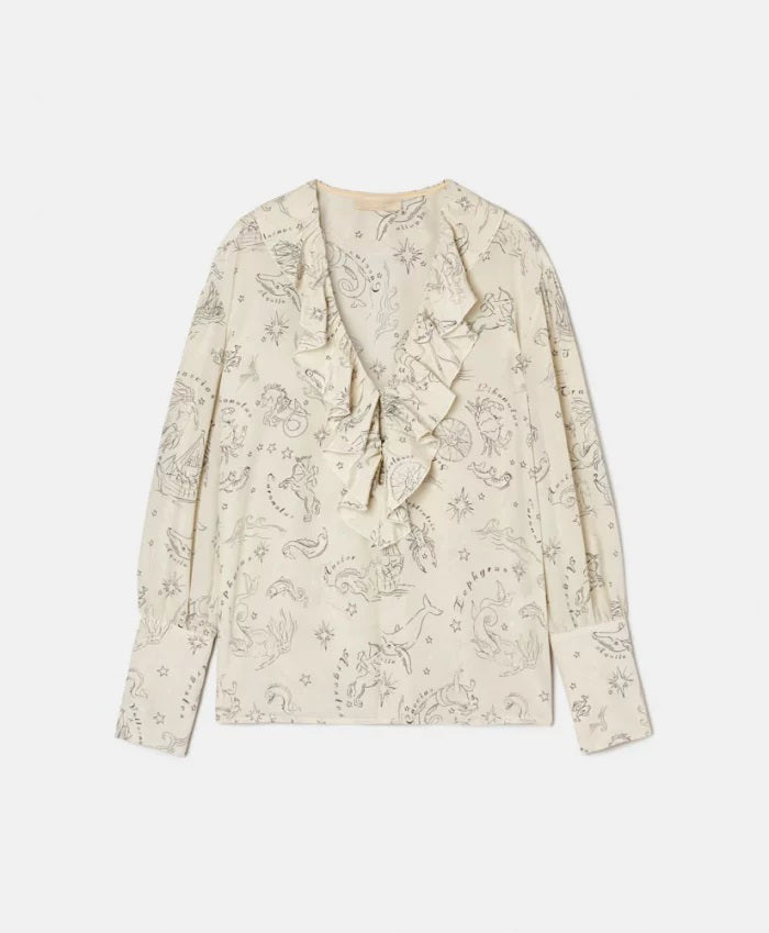 A woman with long brown hair stands outdoors, wearing the MOMONI RUFFLED BLOUSE and striped pants. She looks over her shoulder, with clay jugs and a wooden table in the background against a white wall.