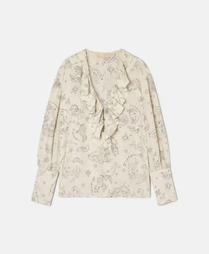 A woman with long brown hair stands outdoors, wearing the MOMONI RUFFLED BLOUSE and striped pants. She looks over her shoulder, with clay jugs and a wooden table in the background against a white wall.