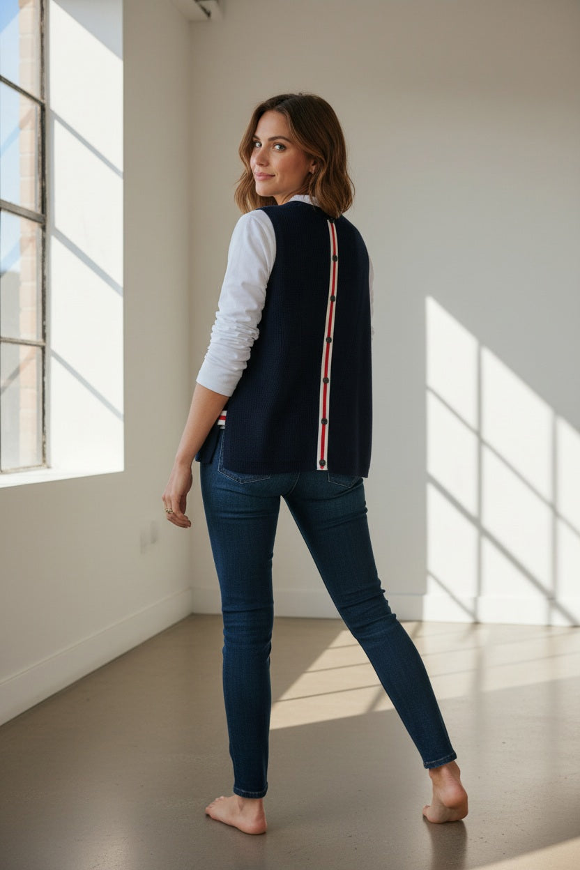 A woman stands barefoot indoors, wearing jeans, a white long-sleeve shirt, and the MERSEA Camden Vest in Navy Cherry White—an oversized sweater vest featuring a red and white striped detail down the back. She glances over her shoulder with a smile in a sunlit room.