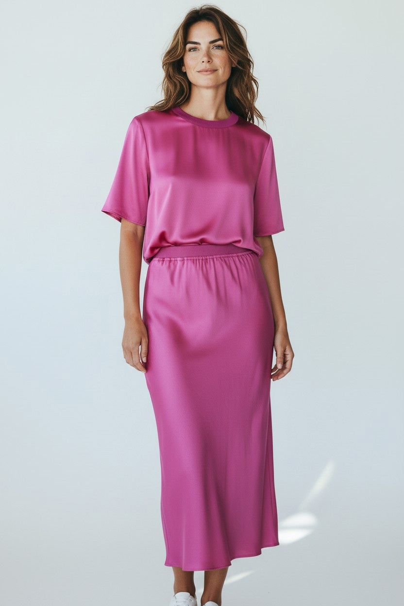 A woman with loose wavy hair and a slight smile poses against a light background wearing the ROSE & CROWN - SATIN TEE TOP in shiny fuchsia, paired with a matching ankle-length satin skirt.