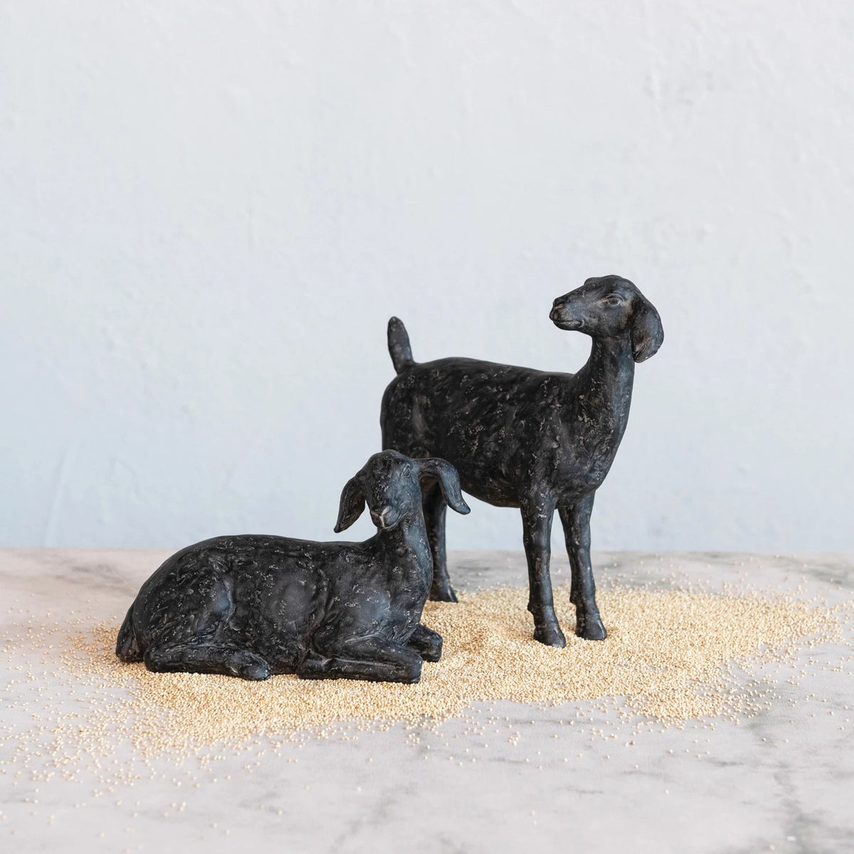 Two small black sculptures of goats from CREATIVE COOP&#39;s RESIN GOAT WITH CAST IRON FINISH collection placed on a light-colored surface create the perfect rustic accent. One goat is standing while the other is lying down, surrounded by scattered grains. The background features a soft, light blue-gray wall, making this scene ideal for animal enthusiasts and elegant goat decor.