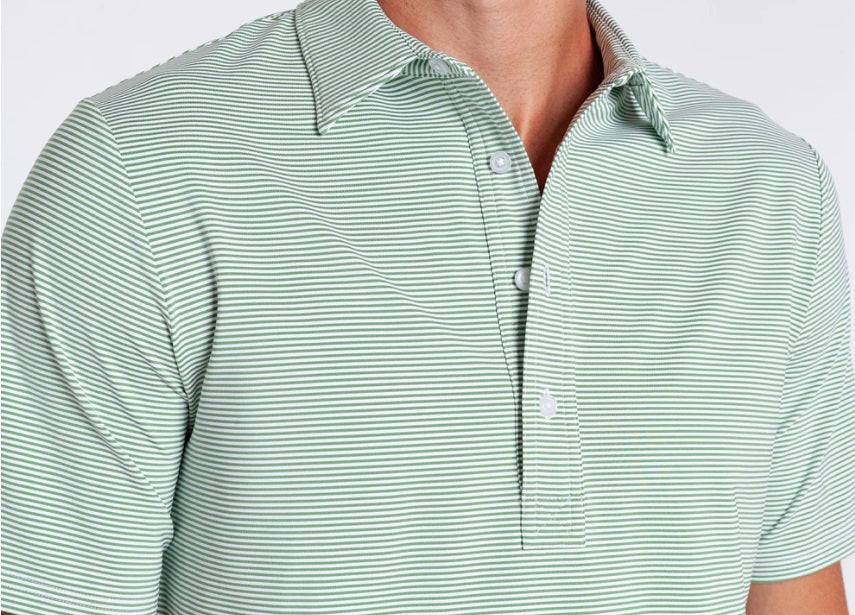A man wears the CRIQUET Performance Sport Range Polo in Hobby Stripe - Gator, featuring light green and white stripes, a collar, and buttons, standing with one hand in his pocket against a plain background.
