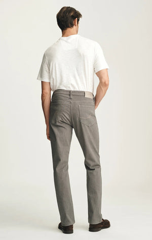 A man stands against a plain background wearing a dark brown sweater layered over a light collared shirt, paired with 34 HERITAGE - COURAGE STRAIGHT LEG PANTS IN STONE DIAGONAL by 34 HERITAGE, and brown shoes. One hand rests in his pocket and he maintains a neutral expression.