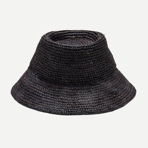 A woman with long, wavy blonde hair wears a sleeveless white top and the WYETH HATS - TALI Raffia Bucket Hat in Midnight by WYETH, featuring an adjustable fit, shown in profile against a plain light background.