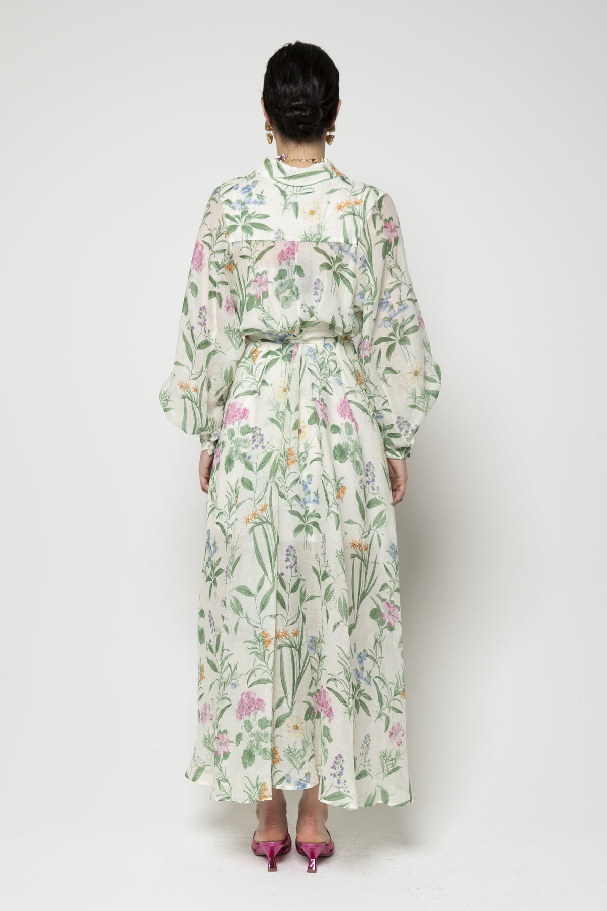 Against a plain background, an individual wears the LAVI COUTURE - ARTEMISIA GREEN FLORAL DRESS, showcasing its luxurious pastel botanical print. They pair it with a colorful necklace and earrings, pink strappy sandals, and neat hair for an elegant look.