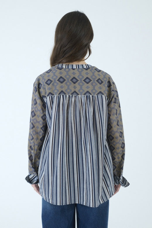 A person with long brown hair, seen from behind, wears the STORIATIPIC MARGOT BLOUSE IN BLUE by STORIATIPIC—a patterned cotton shirt featuring a mix of vertical stripes and diamond shapes in blue and beige tones—paired with blue jeans. The blouse is suitable for a gentle machine wash at 30 degrees.