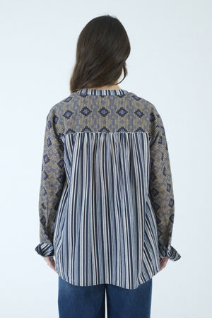A person with long brown hair, seen from behind, wears the STORIATIPIC MARGOT BLOUSE IN BLUE by STORIATIPIC—a patterned cotton shirt featuring a mix of vertical stripes and diamond shapes in blue and beige tones—paired with blue jeans. The blouse is suitable for a gentle machine wash at 30 degrees.