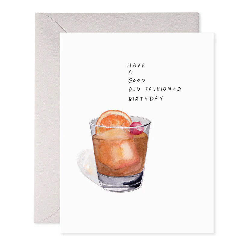 The OLD FASHIONED BIRTHDAY CARD by E.FRANCES PAPER features a beautifully crafted watercolor design of an Old Fashioned cocktail, complete with an orange slice and cherry garnish. The card includes the message, "Have a good old fashioned birthday," and is accompanied by a gray envelope, all printed on luxurious paper.