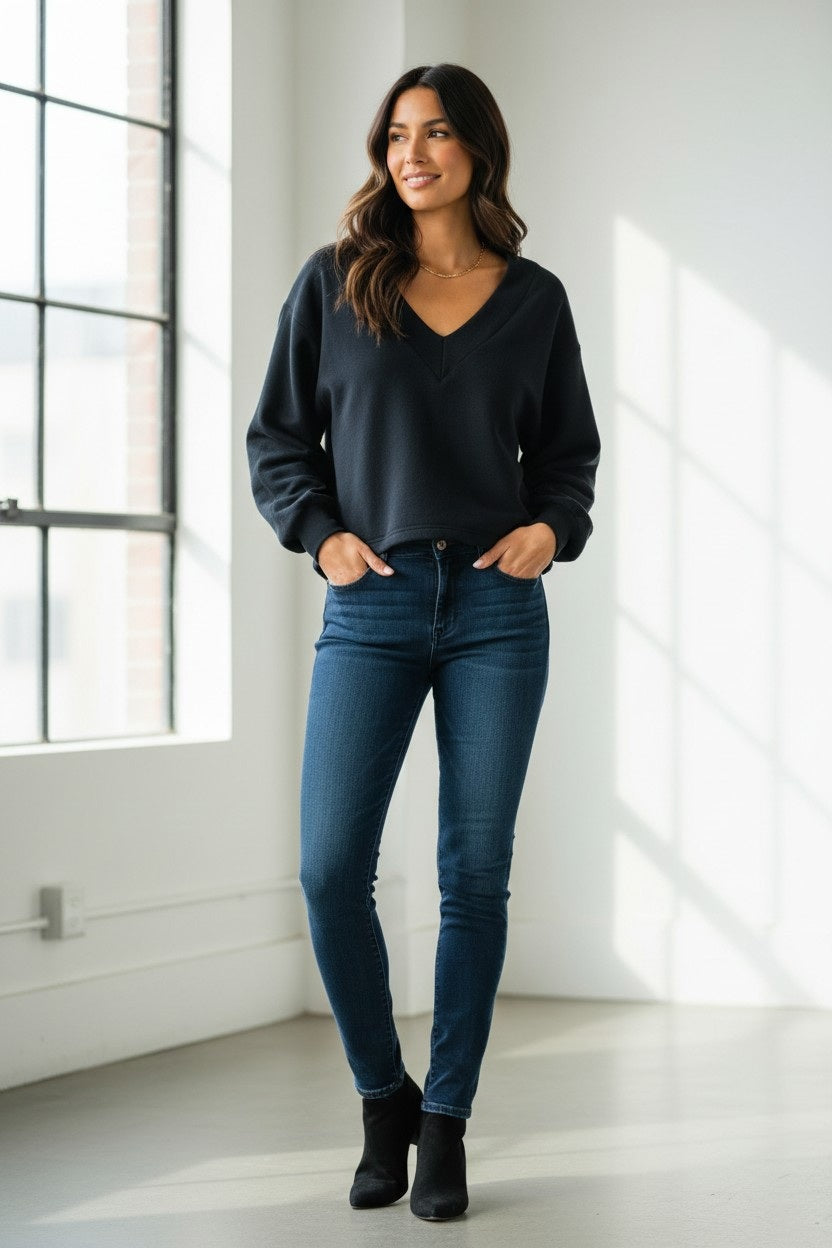 A barefoot woman wearing black sweatpants and the KMJ - TUCKER V NECK SWEATSHIRT IN BLACK by KMJ leans against a cream-colored wall outdoors, gazing to the side. Two large windows are visible behind her, with black sandals on the ground nearby.