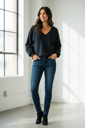 A barefoot woman wearing black sweatpants and the KMJ - TUCKER V NECK SWEATSHIRT IN BLACK by KMJ leans against a cream-colored wall outdoors, gazing to the side. Two large windows are visible behind her, with black sandals on the ground nearby.
