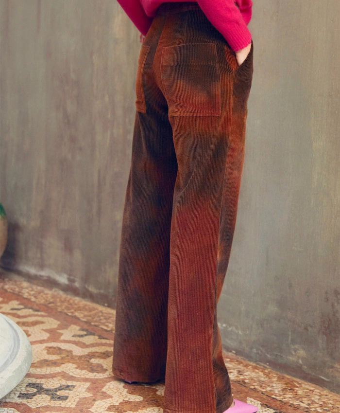 A person with long dark hair stands against a muted indoor wall, wearing a bright pink sweater, MOMONI - ISMAELTROUSER tie-dyed corduroy pants, and pink shoes. They have their hands in the patch pockets and accessorize with two long necklaces. An urn and a small water pool are placed beside them.
