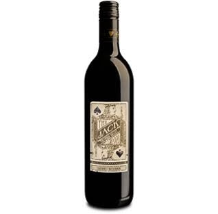 A bottle of SAVIAH ‘THE JACK’ CABERNET 2022 from WINE: OCEAN SOUTH IMPORTS and a filled wine glass rest on a white table, with shelves of assorted wine bottles in the background.