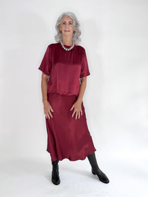 A shiny ROSE & CROWN - SATIN CREW NECK TEE in rose gold hangs on a white hanger against a plain white background, shown from the back with short sleeves.