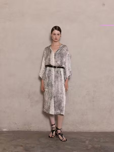 A woman stands against a beige wall, wearing the NUDE - ANIMALIER DRESS by NUDE. The dress features a loose, kaftan-inspired silhouette with black-and-white animal print, styled with wide-leg pants. Her hair is straight and pulled back, with a neutral expression.