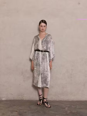 A woman stands against a beige wall, wearing the NUDE - ANIMALIER DRESS by NUDE. The dress features a loose, kaftan-inspired silhouette with black-and-white animal print, styled with wide-leg pants. Her hair is straight and pulled back, with a neutral expression.