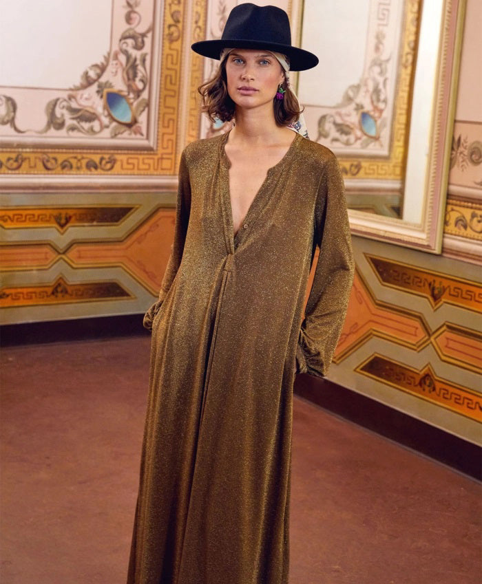 A person stands in a lavishly decorated room, wearing the shimmering long MOMONI - AMEDE DRESS IN LUREX JERSEY, made in Italy by MOMONI, paired with a black wide-brimmed hat adorned with a colorful band. Their hands are in their pockets, and there's a large mirror reflecting part of the ornate wall design.