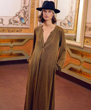 A person stands in a lavishly decorated room, wearing the shimmering long MOMONI - AMEDE DRESS IN LUREX JERSEY, made in Italy by MOMONI, paired with a black wide-brimmed hat adorned with a colorful band. Their hands are in their pockets, and there's a large mirror reflecting part of the ornate wall design.