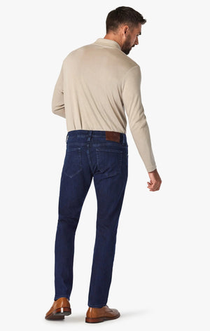 A man stands with his back to the camera, wearing a beige long-sleeve shirt, 34 HERITAGE - COURAGE STRAIGHT LEG JEAN IN DARK MIDNIGHT BRUSHED URBAN WASH, and brown shoes against a plain white background.