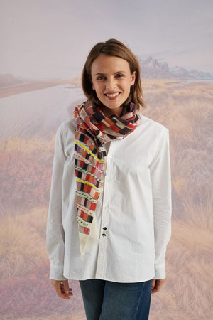 A woman with light brown hair wears a cream shirt and the STORIATIPIC - ANOUK WOOL SCARF, an elegant hand-embroidered accessory by STORIATIPIC, wrapped around her neck. Mountains and plains form a soft, scenic backdrop.