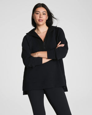 A woman with long dark hair stands confidently with arms crossed, wearing the SPANX - AirEssentials Brushed Half Zip Tunic Top in Black and matching leggings against a plain light gray background, showcasing stylish loungewear.