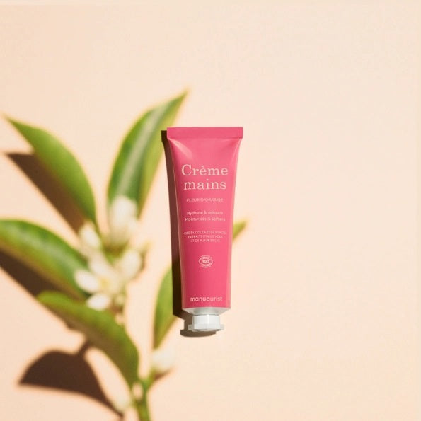 Close-up of two hands with light skin; one is gently applying MANUCURIST - COSMOS ORGANIC HAND CREAM, ORANGE BLOSSOM by MANUCURIST INC to the back of the other against a neutral beige background. Ideal for hand and nail care, this organic moisturizer is formulated with natural ingredients to leave fingers nourished. The nails are perfectly groomed and finished with a subtle pale nude polish for an elegant touch.