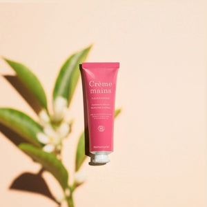 Close-up of two hands with light skin; one is gently applying MANUCURIST - COSMOS ORGANIC HAND CREAM, ORANGE BLOSSOM by MANUCURIST INC to the back of the other against a neutral beige background. Ideal for hand and nail care, this organic moisturizer is formulated with natural ingredients to leave fingers nourished. The nails are perfectly groomed and finished with a subtle pale nude polish for an elegant touch.