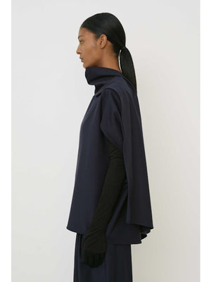 A woman with slicked-back hair and a neutral expression wears the MEIMEIJ Draped Cape Effect Top in navy over black long sleeves, paired with a matching navy skirt, set against a plain white background.