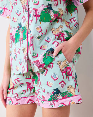 A woman with long blonde hair smiles as she stands against a plain white background, wearing the PRINTFRESH - MAHJONG MENAGERIE PAJAMA SHORT SET by PRINTFRESH, a matching short-sleeve button-up pajama top and shorts hand screen-printed with colorful cat and chair patterns.