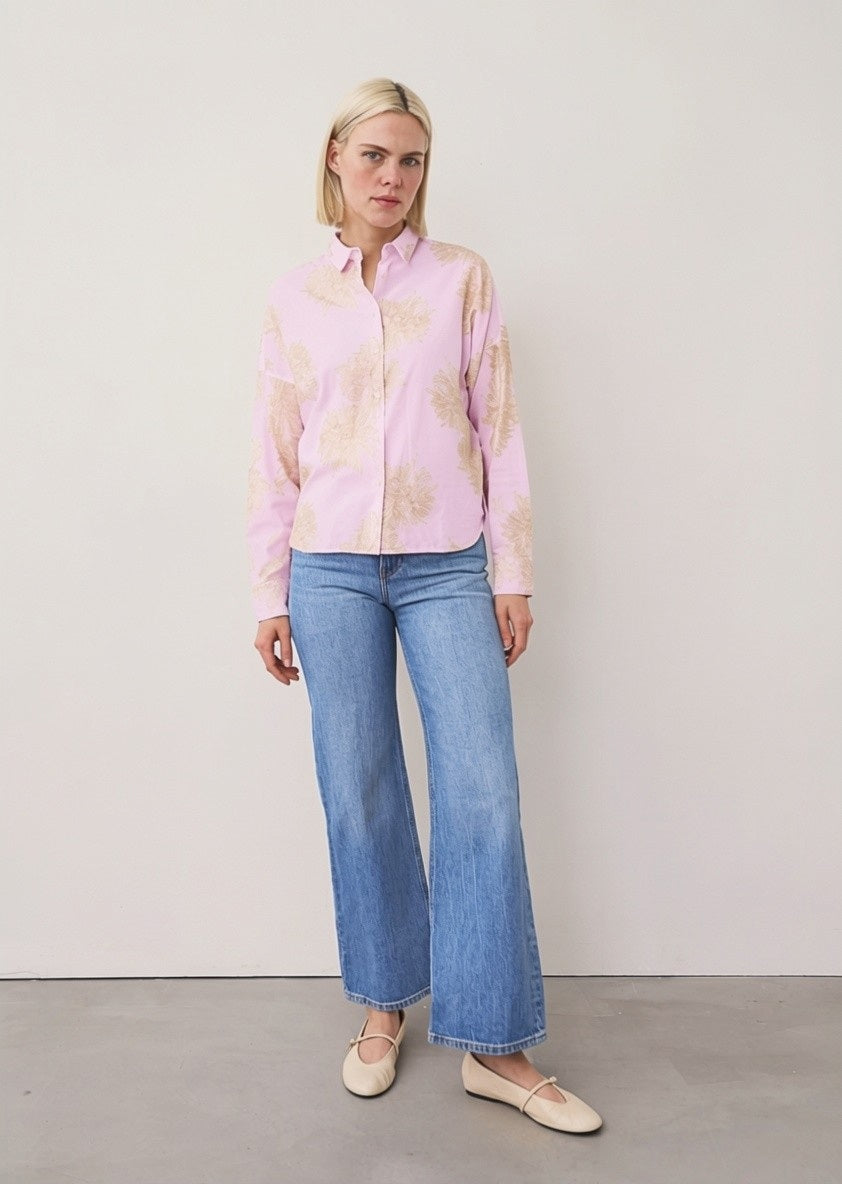A woman with straight blonde hair and a neutral expression stands against a plain wall, wearing the LUCY BLOUSE WITH EMBROIDERED FLOWER by 0039 ITALY, blue wide-leg jeans with relaxed tailoring, and beige ballet flats.