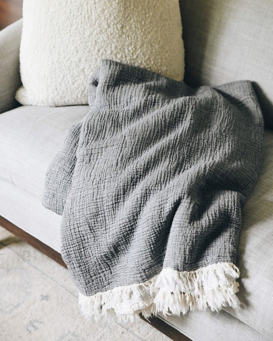 The MERSEA CLOUD GAUZE BLANKET is a folded, textured gray cotton throw with a ribbed pattern and off-white fringe along the edges, shown against a plain light background.