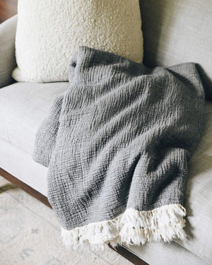 The MERSEA CLOUD GAUZE BLANKET is a folded, textured gray cotton throw with a ribbed pattern and off-white fringe along the edges, shown against a plain light background.