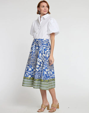 A person is modeling a white blouse with puffed sleeves and the HINSON WU - GLORIA SKIRT IN CERAMIC TILE PRINT, featuring a blue and white pattern with green and yellow striped accents at the hem. They are wearing beige open-toed, block-heeled sandals.