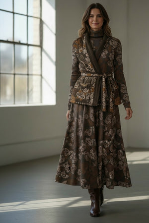 A woman stands indoors by a window, smiling softly with wavy hair. She wears the FRANCO FERRARI - REVERSIBLE KIMONO JACKET, which features a patterned velvet design and a subtle kimono silhouette, paired with a black top, black pants, and black loafers.