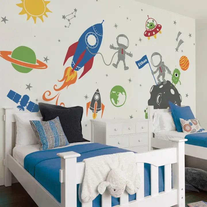 Children's bedroom with two white beds featuring blue and striped bedding. The wall is decorated with SIMPLE SHAPES - OUTER SPACE REMOVABLE WALL DECALS by SIMPLE SHAPES, including rockets, astronauts, planets, stars, and aliens. The wall also has a constellation and sun illustration.